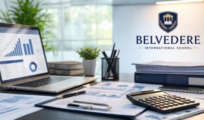 A modern, well-lit office workspace with a laptop displaying financial charts, accounting documents spread across a desk, a calculator, pen holder, and stacked files. On the wall, a clean logo reads “Belvedere International School,” giving a professional corporate atmosphere.